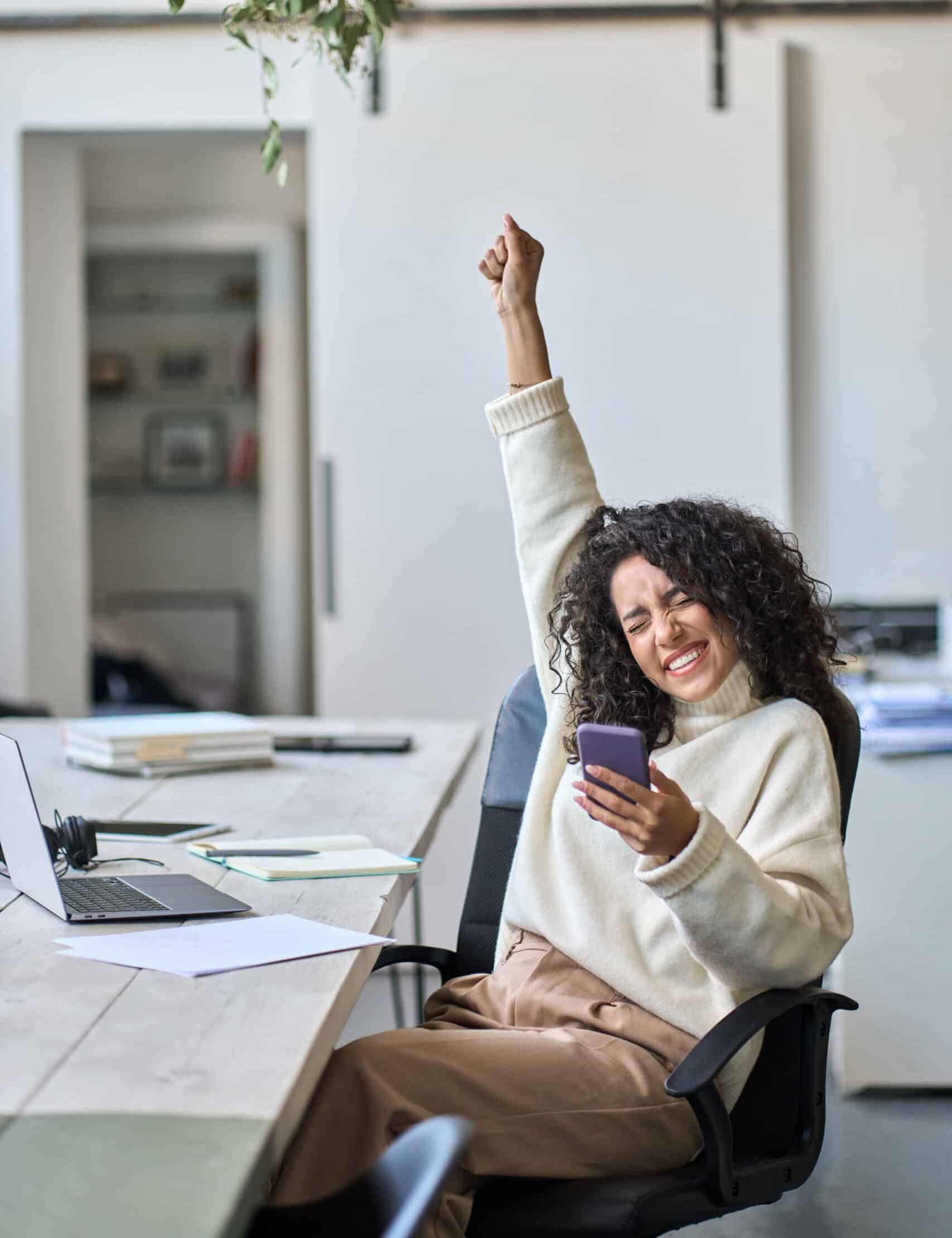 Woman holding phone, celebrating at a desk.