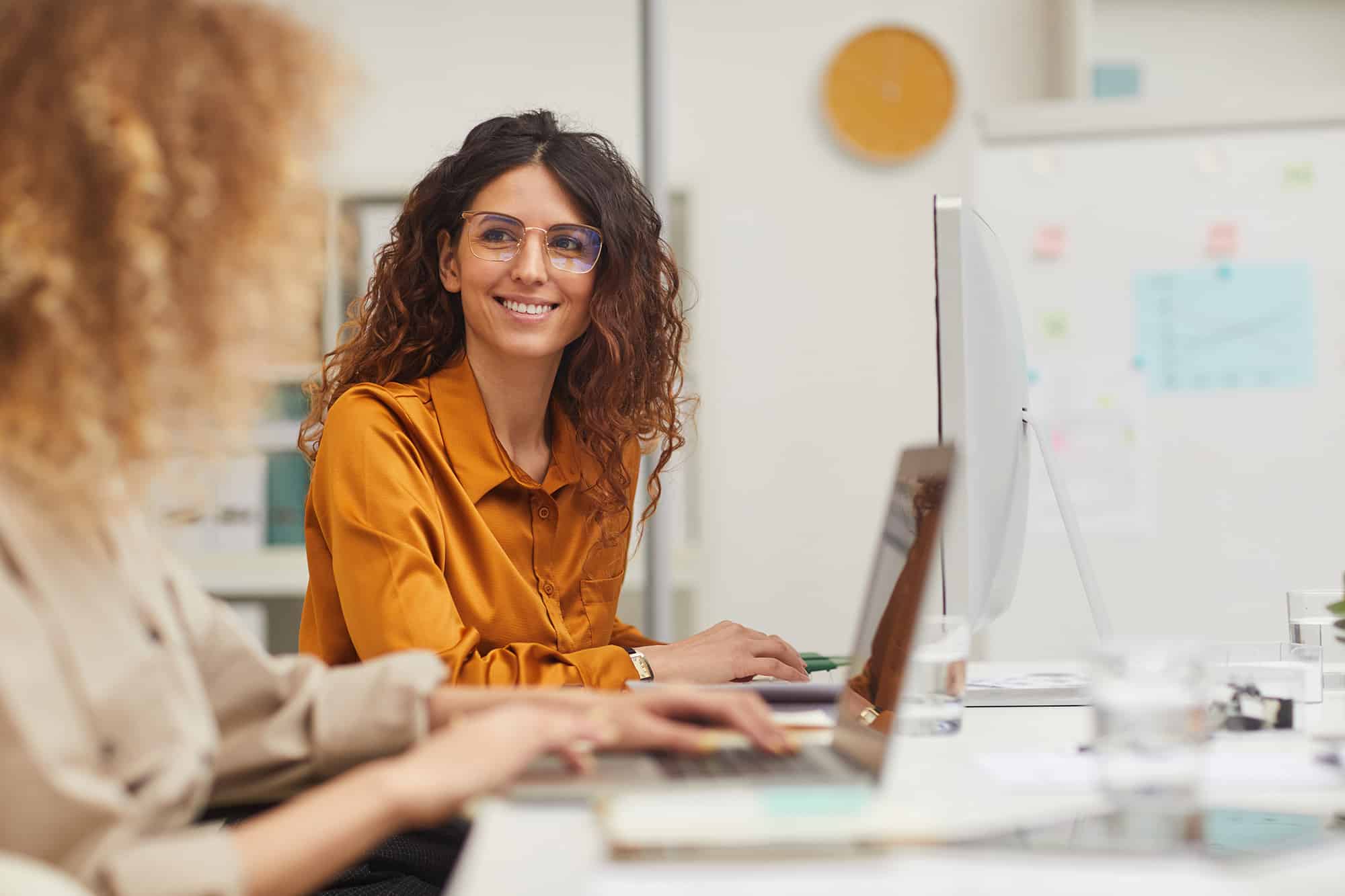A woman with curly hair and a yellow shirt collaborates with a co-worker on a compter.